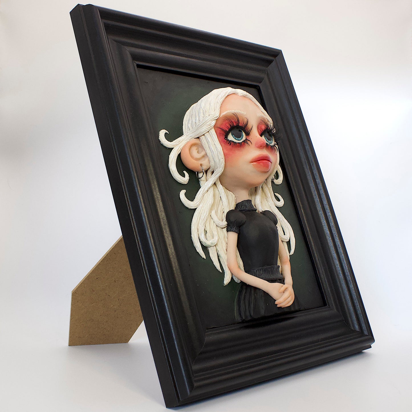 A polymer clay sculpture positioned in a black wood photo frame. Featuring a haunting girl with blond hair and a black Victorian style gothic dress, set against a green black gradient background.