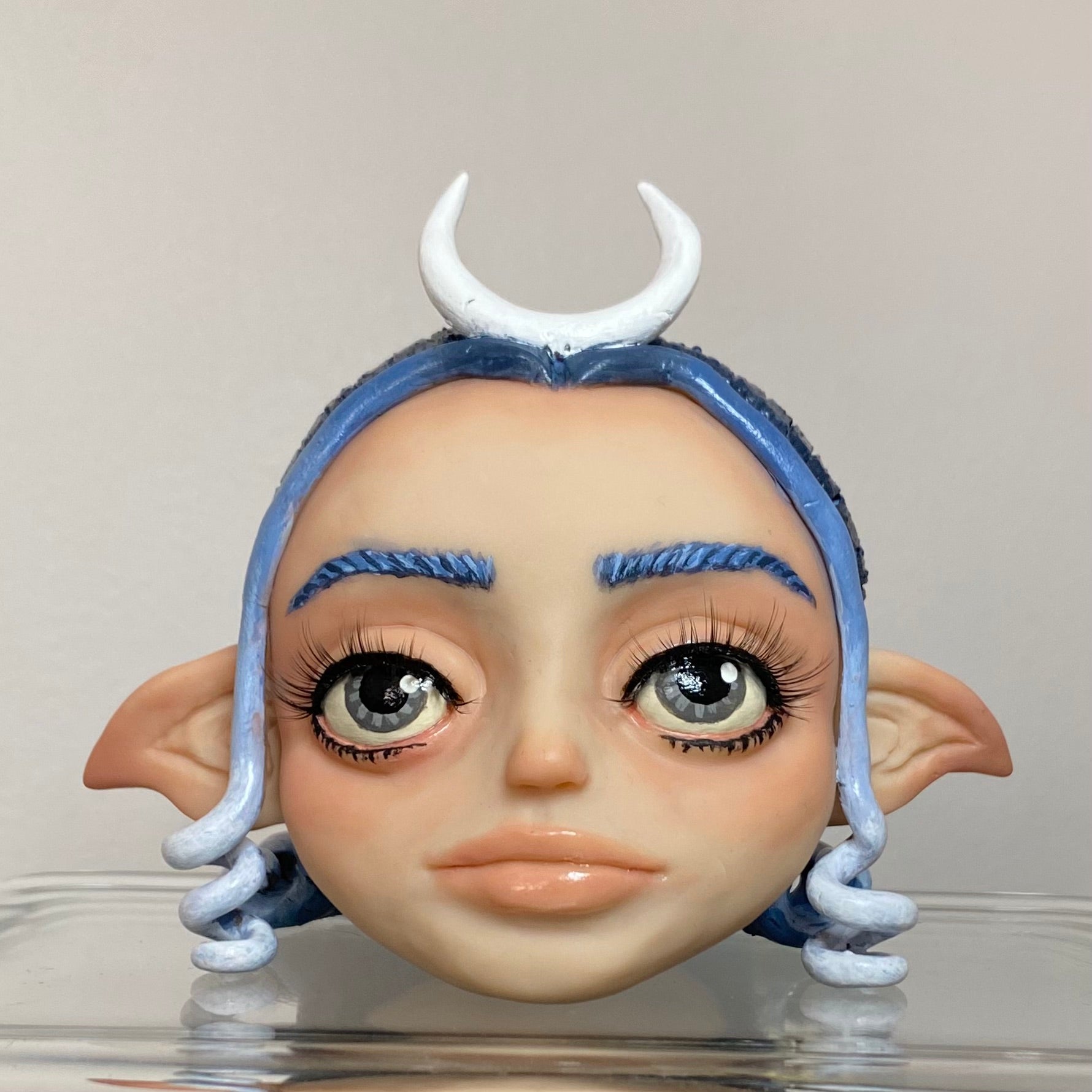 Polymer clay sculpture of a moon elf head, featuring a moon shaped crown, hair in a gradient from dark blue to light blue. 