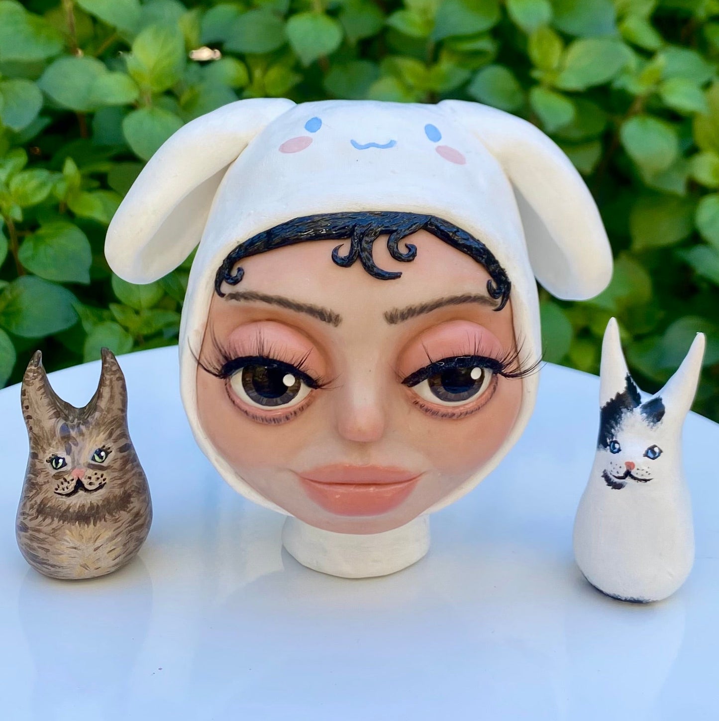 A polymer clay art sculpture of Vanilla Mace's head in a cinamoroll beanie and her two cats, Miso and Chai.