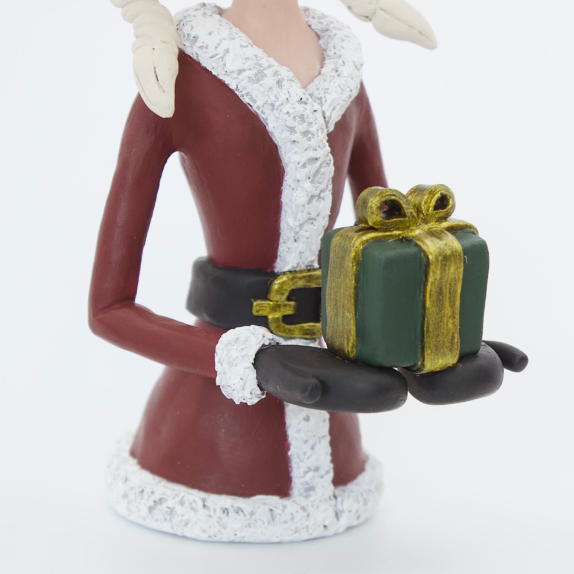 A polymer clay sculpture featuring a young mrs claus with blond hair holding a Christmas present gift box, wearing a Santa hat and suit. 