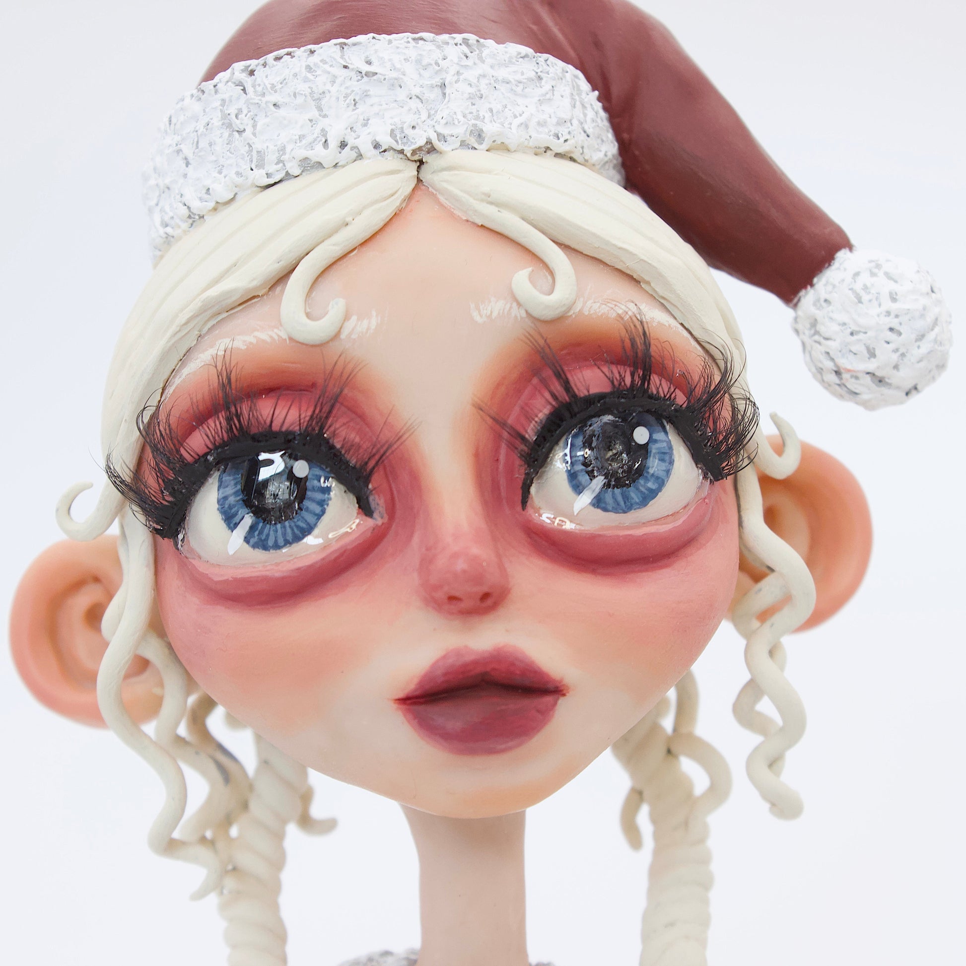 A polymer clay sculpture featuring a young mrs claus with blond hair holding a Christmas present gift box, wearing a Santa hat and suit. 