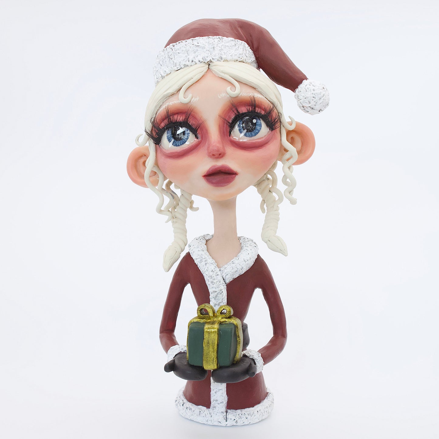 A polymer clay sculpture featuring a young mrs claus with blond hair holding a Christmas present gift box, wearing a Santa hat and suit. 