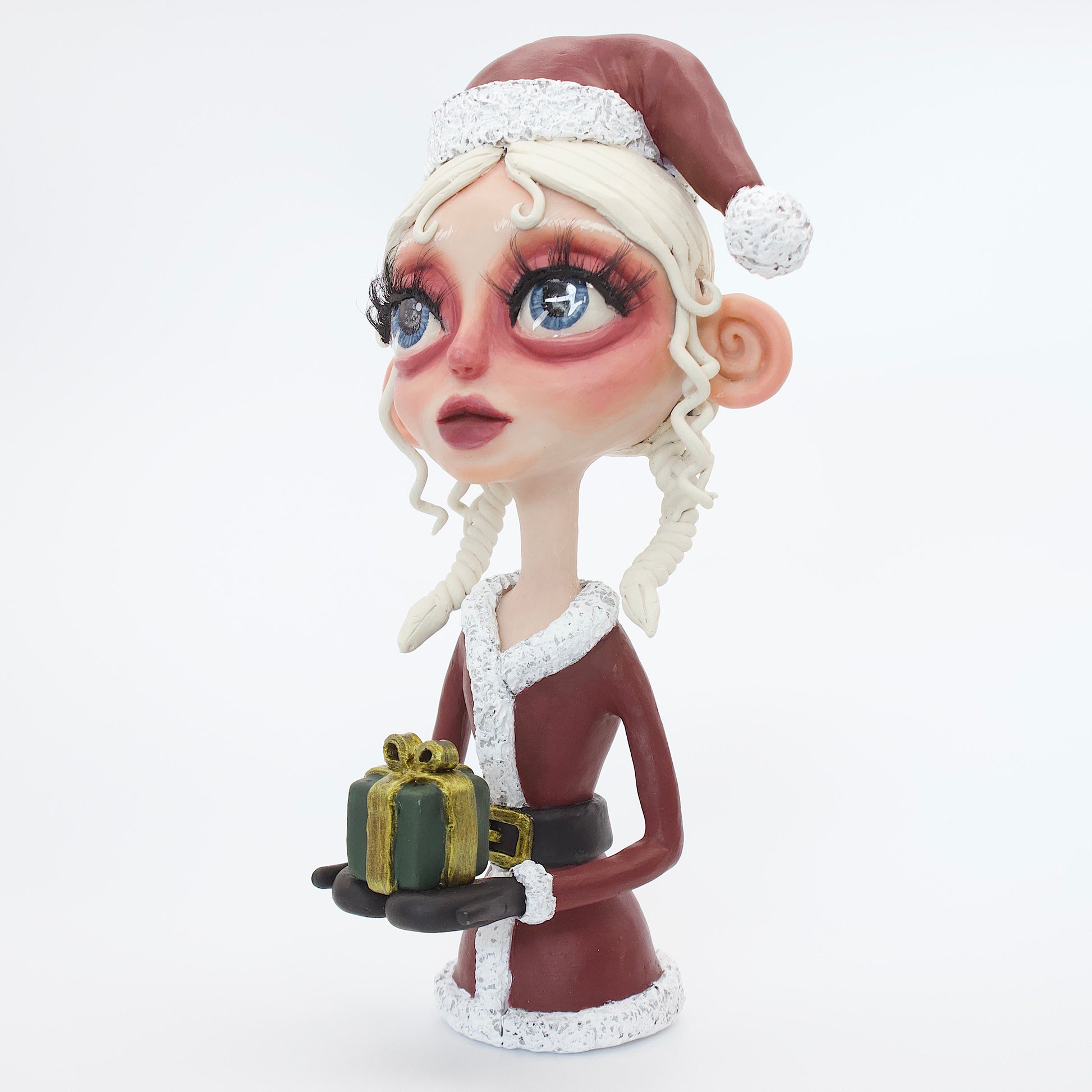 A polymer clay sculpture featuring a young mrs claus with blond hair holding a Christmas present gift box, wearing a Santa hat and suit. 