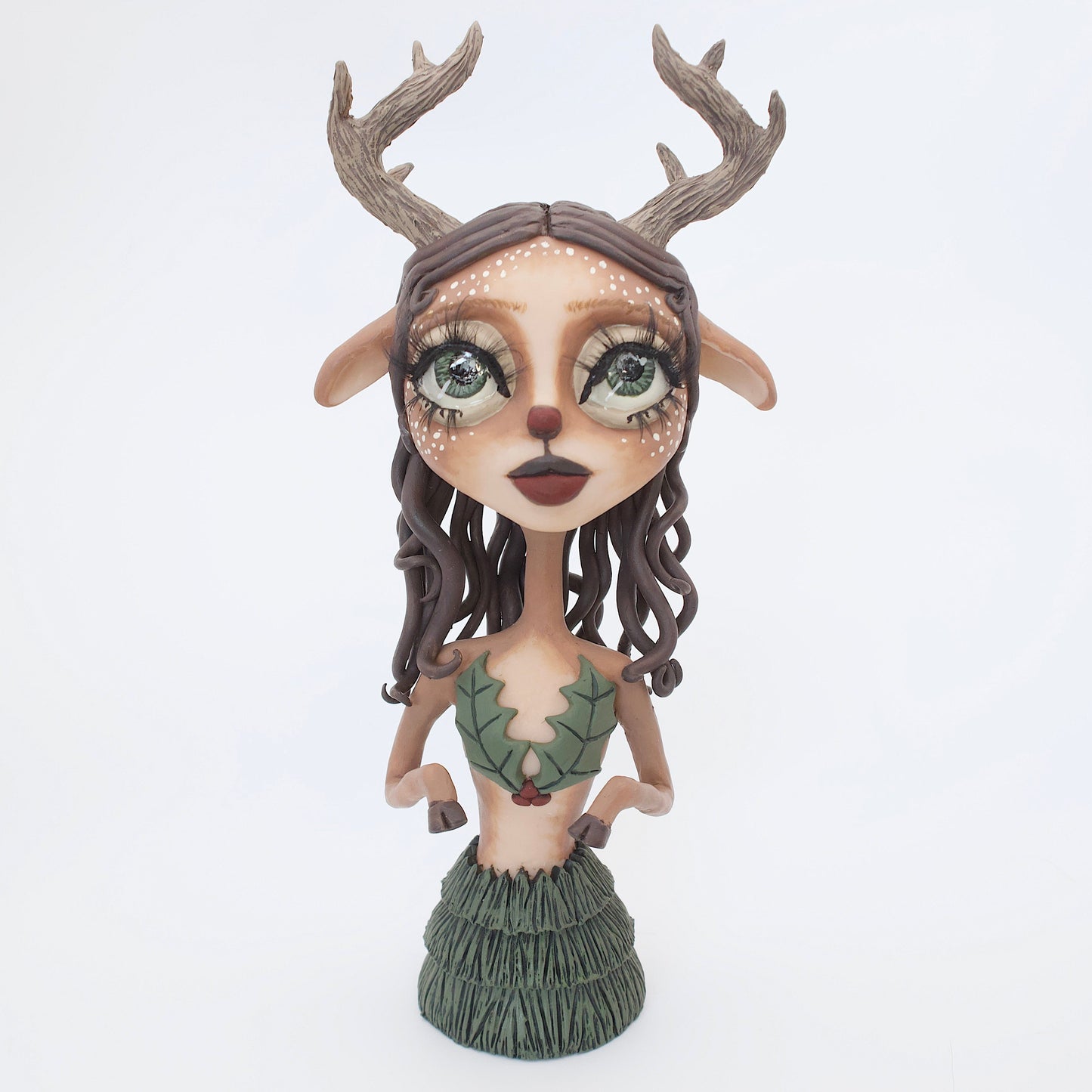 A polymer clay sculpture of a reindeer human hybrid, featuring antlers, a christmas tree skirt, and holly berries and leaves for the top. Also with the iconic Rudolph red nose and deer like ears and hooves.