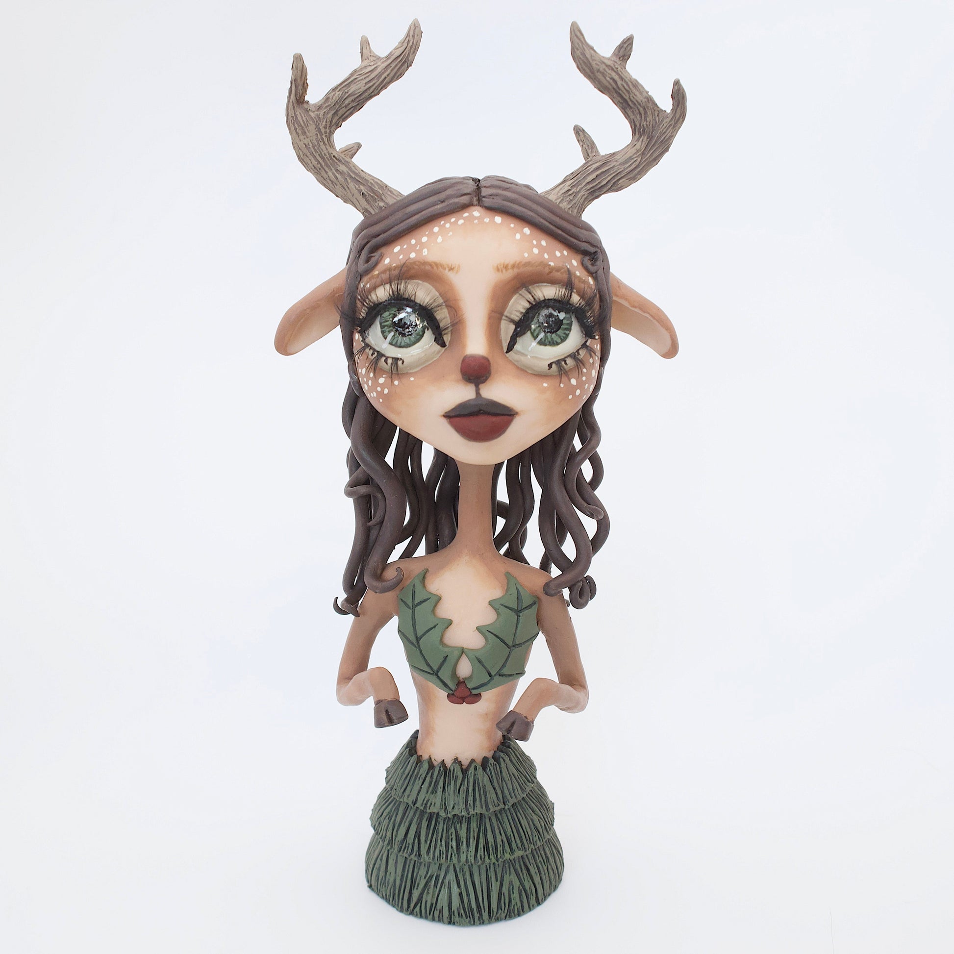 A polymer clay sculpture of a reindeer human hybrid, featuring antlers, a christmas tree skirt, and holly berries and leaves for the top. Also with the iconic Rudolph red nose and deer like ears and hooves.