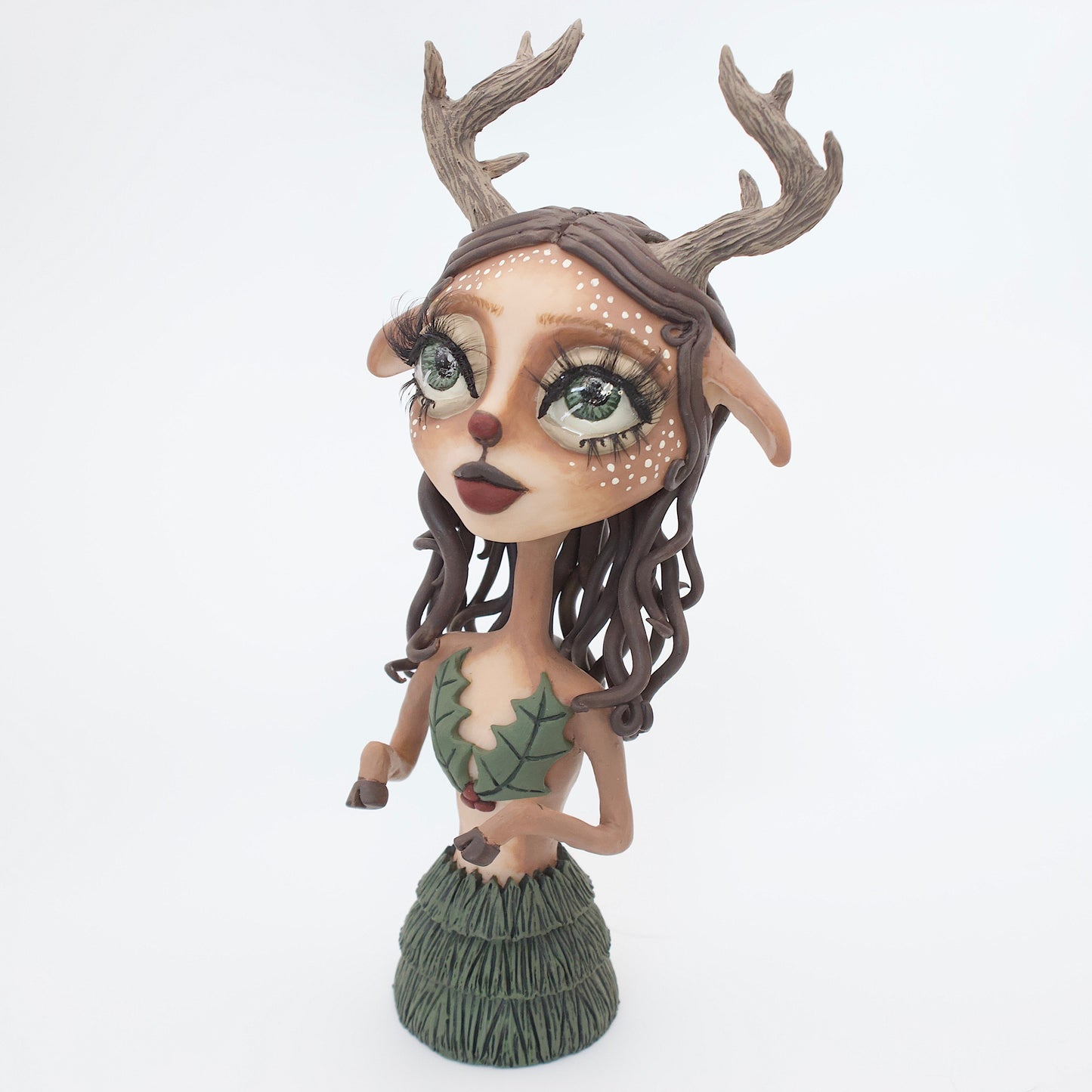 A polymer clay sculpture of a reindeer human hybrid, featuring antlers, a christmas tree skirt, and holly berries and leaves for the top. Also with the iconic Rudolph red nose and deer like ears and hooves.