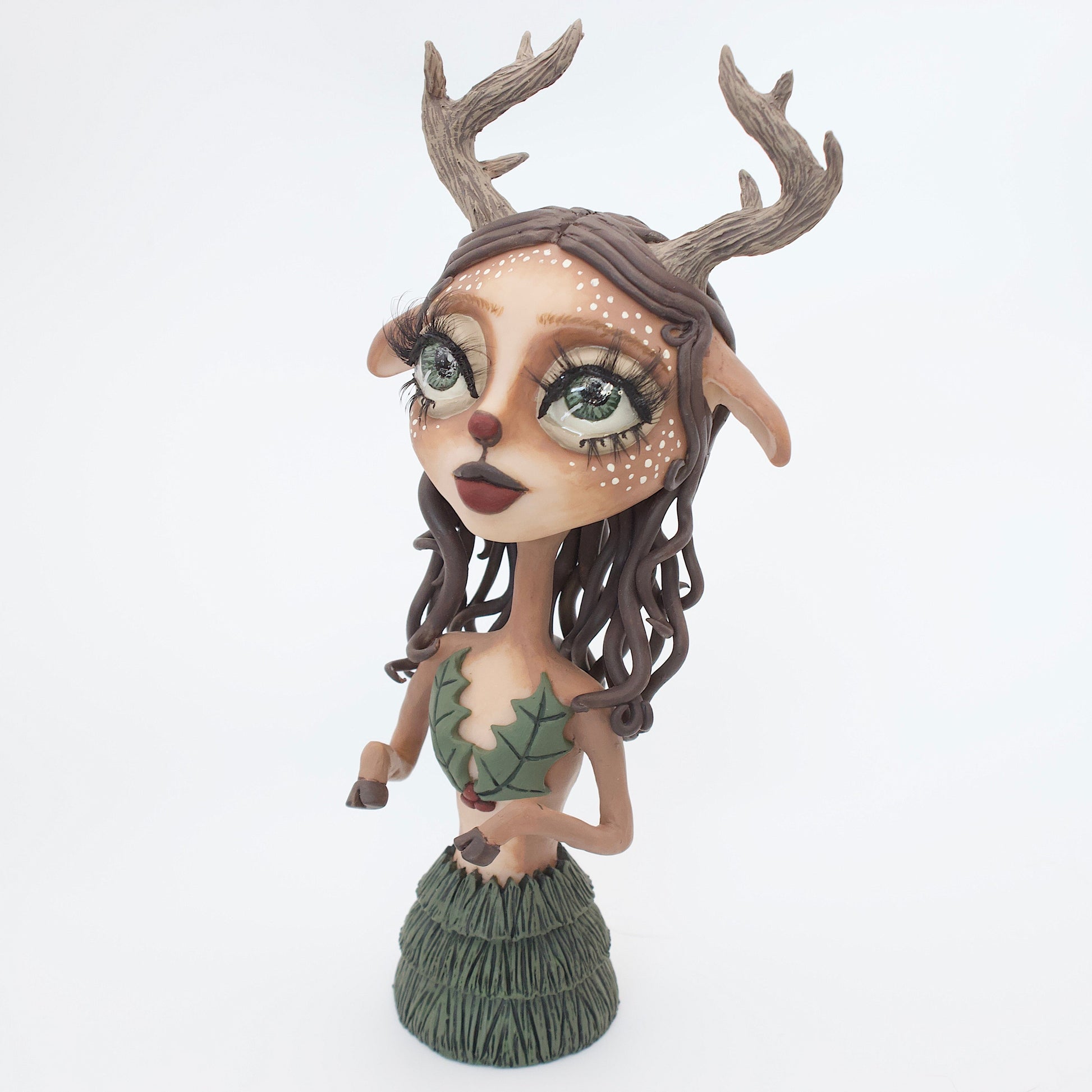 A polymer clay sculpture of a reindeer human hybrid, featuring antlers, a christmas tree skirt, and holly berries and leaves for the top. Also with the iconic Rudolph red nose and deer like ears and hooves.