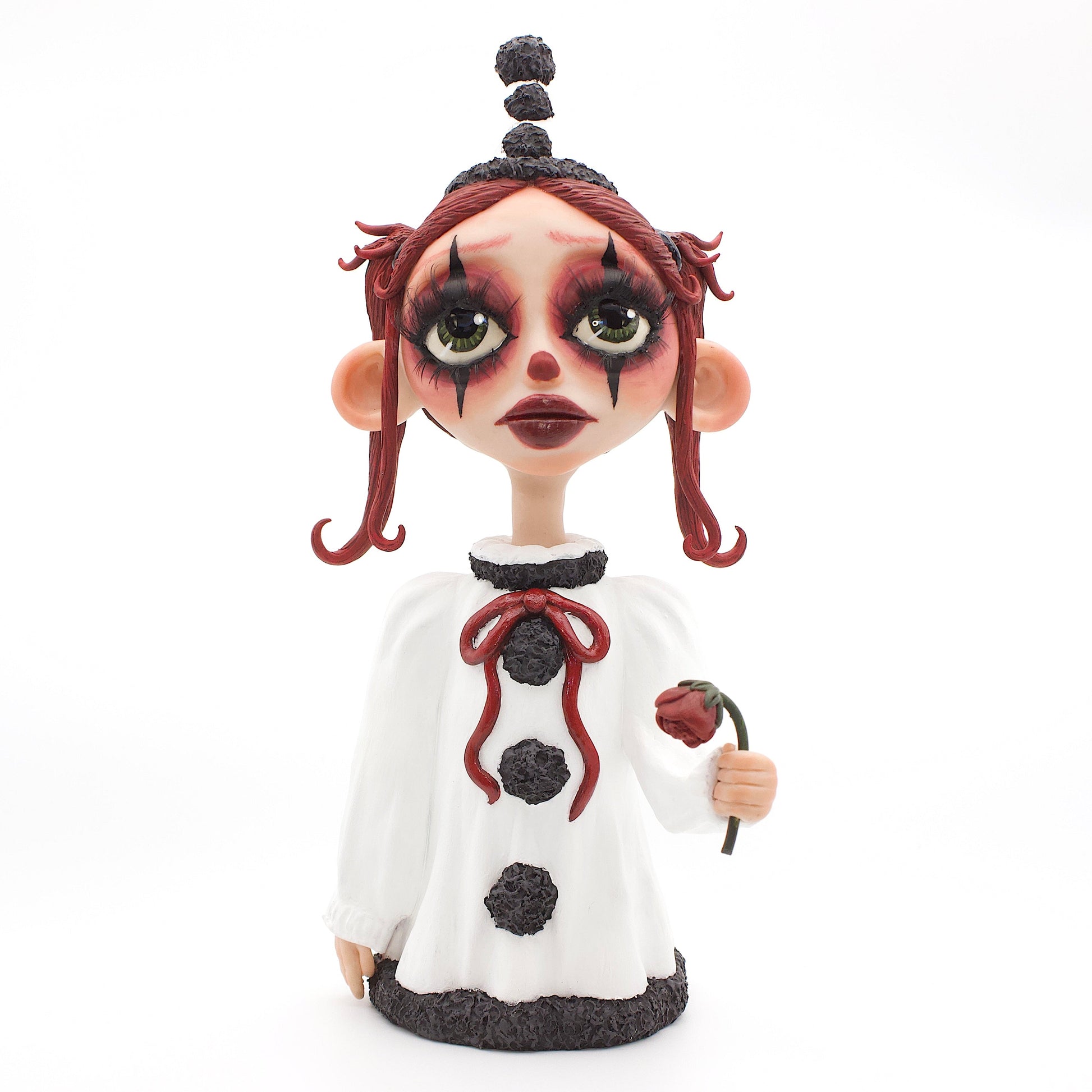 A polymer clay sculpture of a sad pierrot girl clown, holding a drooping rose, wearing the iconic pierrot clown garb in black white and red.