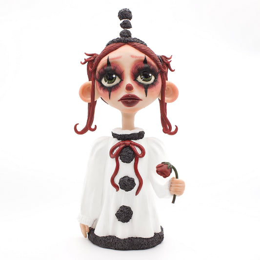A polymer clay sculpture of a sad pierrot girl clown, holding a drooping rose, wearing the iconic pierrot clown garb in black white and red.