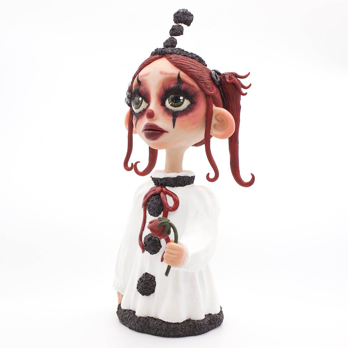 A polymer clay sculpture of a sad pierrot girl clown, holding a drooping rose, wearing the iconic pierrot clown garb in black white and red.
