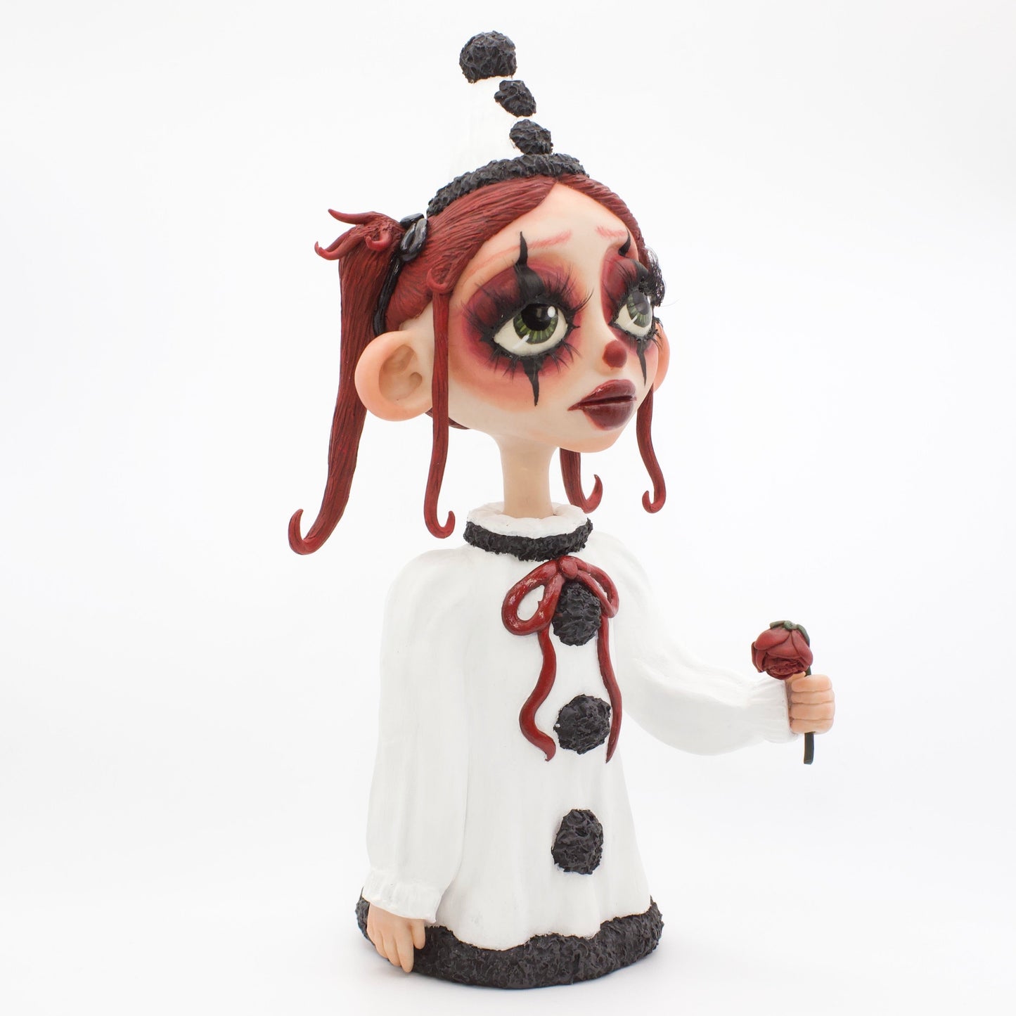 A polymer clay sculpture of a sad pierrot girl clown, holding a drooping rose, wearing the iconic pierrot clown garb in black white and red.