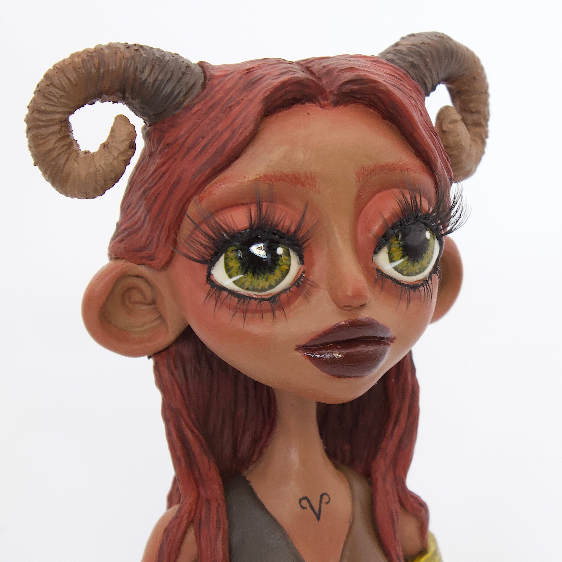 A polymer clay sculpture of the Aries zodiac personified as a woman, featuring red toned browns, long red brown hair and the iconic ram horns.