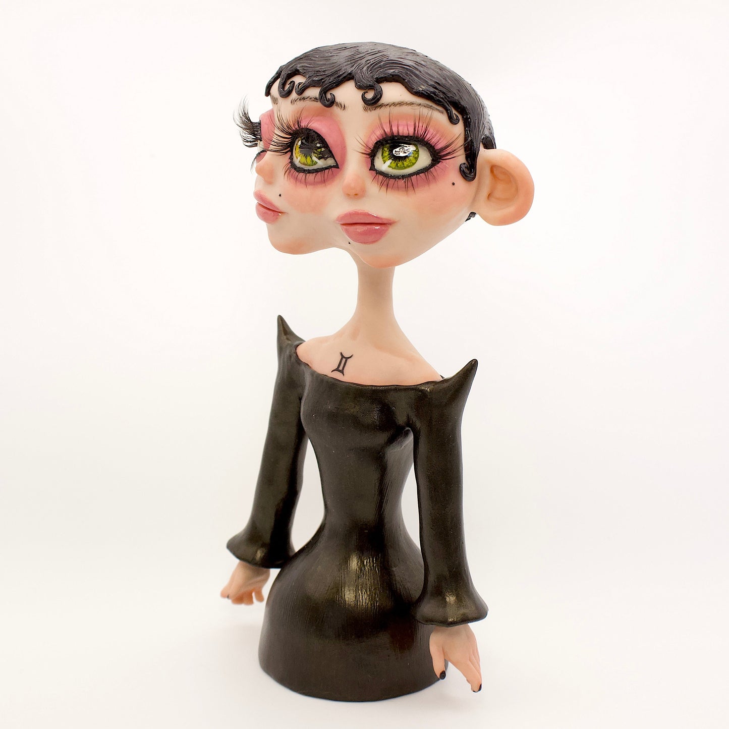 A polymer clay sculpture of the Gemini zodiac personified as a woman, featuring a sleek black dress and two heads fused together, sharing an eye.