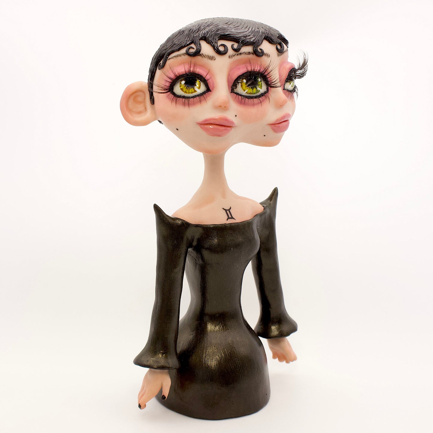 A polymer clay sculpture of the Gemini zodiac personified as a woman, featuring a sleek black dress and two heads fused together, sharing an eye.