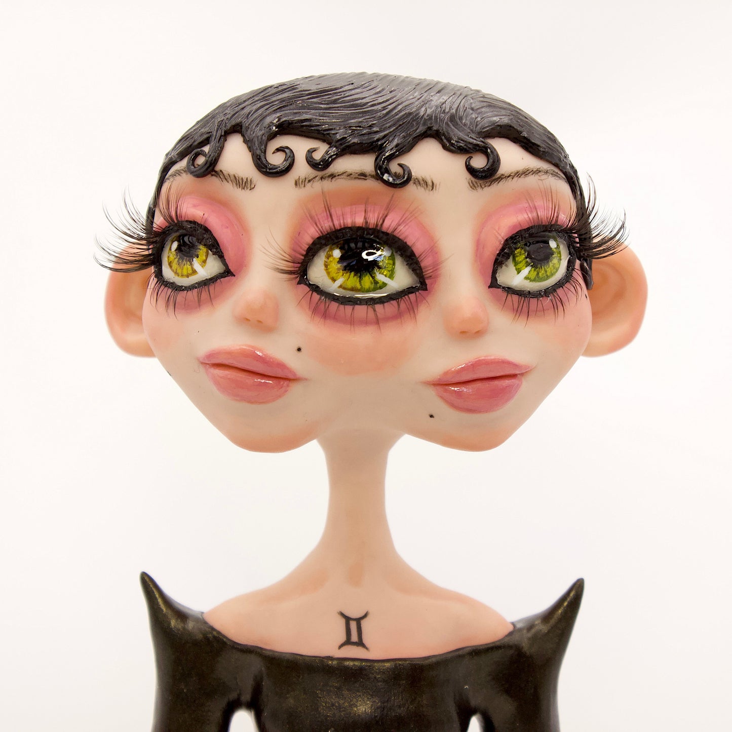 A polymer clay sculpture of the Gemini zodiac personified as a woman, featuring a sleek black dress and two heads fused together, sharing an eye.