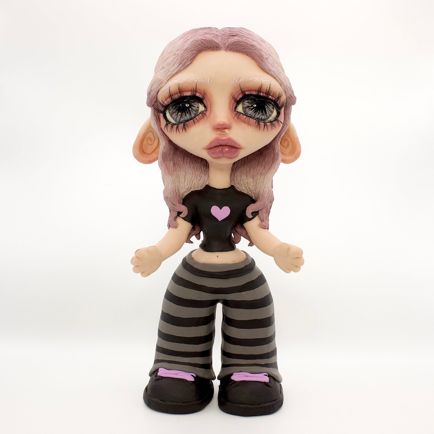 A stylized doll or figurine with large eyes, pale skin, lavender hair, a black top with a pink heart, and black-and-gray striped pants, standing against a white studio backdrop.
