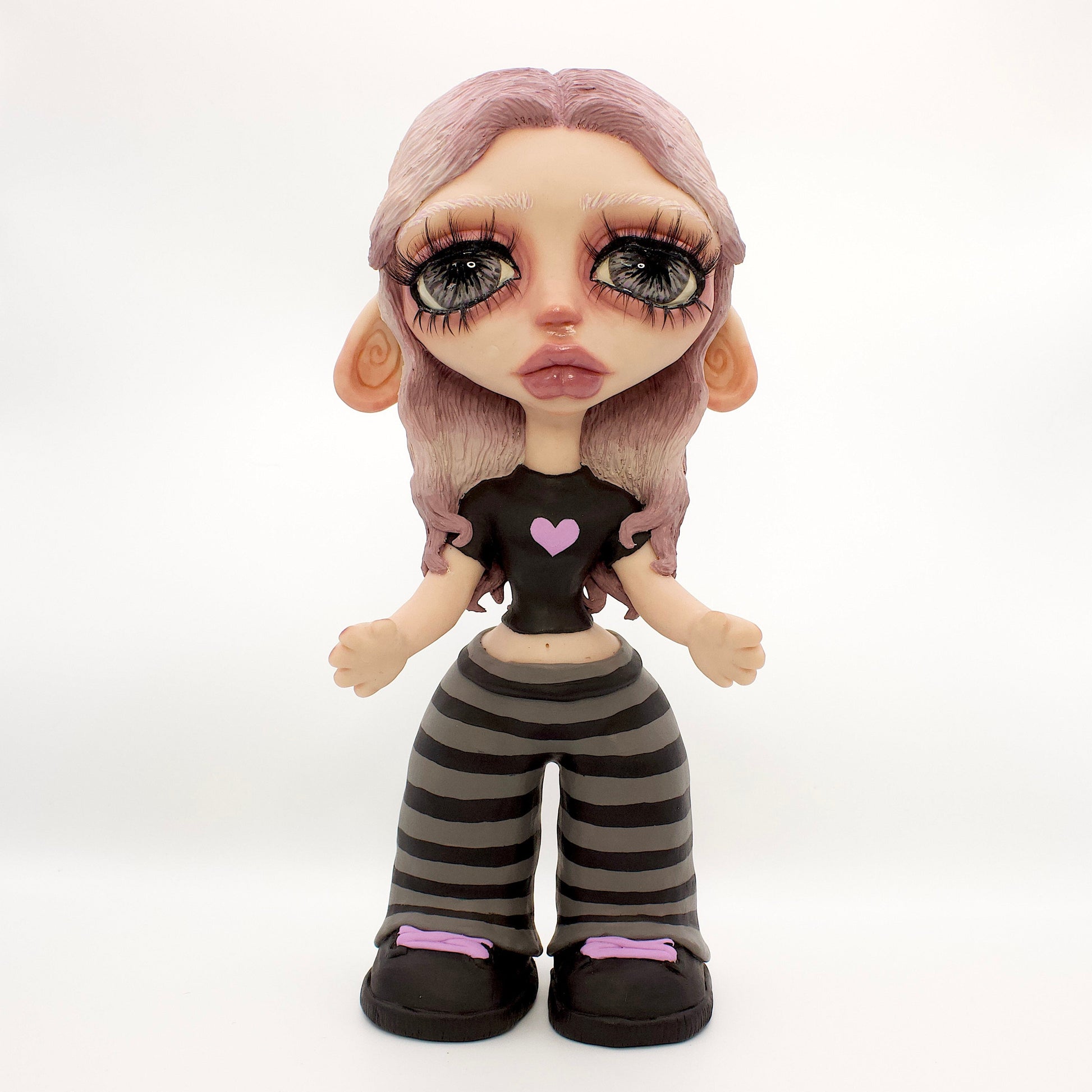 A stylized doll or figurine with large eyes, pale skin, lavender hair, a black top with a pink heart, and black-and-gray striped pants, standing against a white studio backdrop.