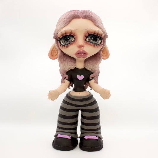 A stylized doll or figurine with large eyes, pale skin, lavender hair, a black top with a pink heart, and black-and-gray striped pants, standing against a white studio backdrop.