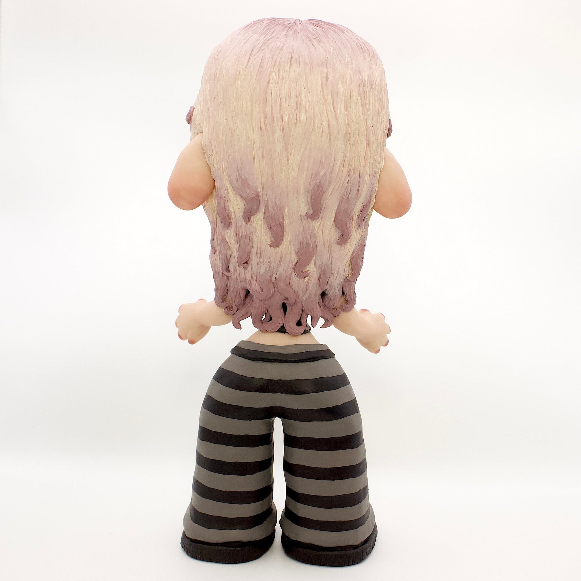 A stylized doll or figurine with large eyes, pale skin, lavender hair, and a black top with a pink heart, paired with black-and-gray striped pants and black shoes, photographed against a white studio backdrop.