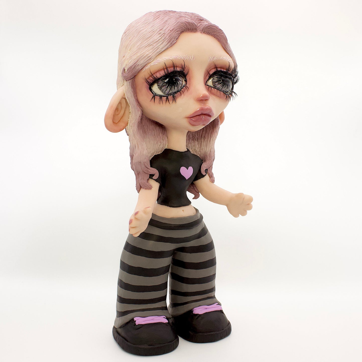 A stylized doll with large eyes, pale skin, lavender hair, a black top with a small pink heart, and gray-and-black striped pants, standing against a white studio backdrop.