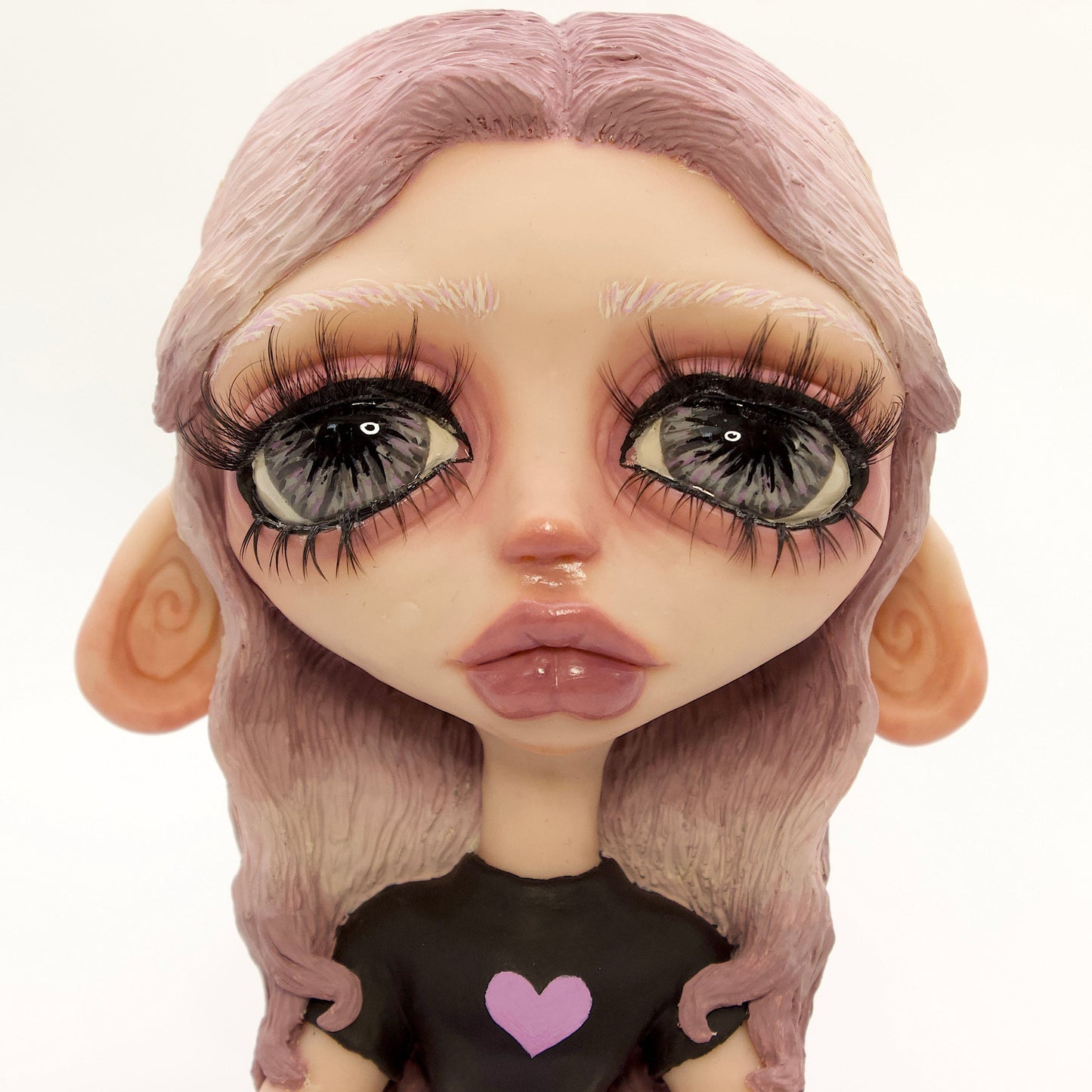 A close-up of a stylized doll or figurine with oversized glossy eyes, pale skin, lavender-pink hair, and a black top with a small pink heart on the chest, photographed against a plain white background.