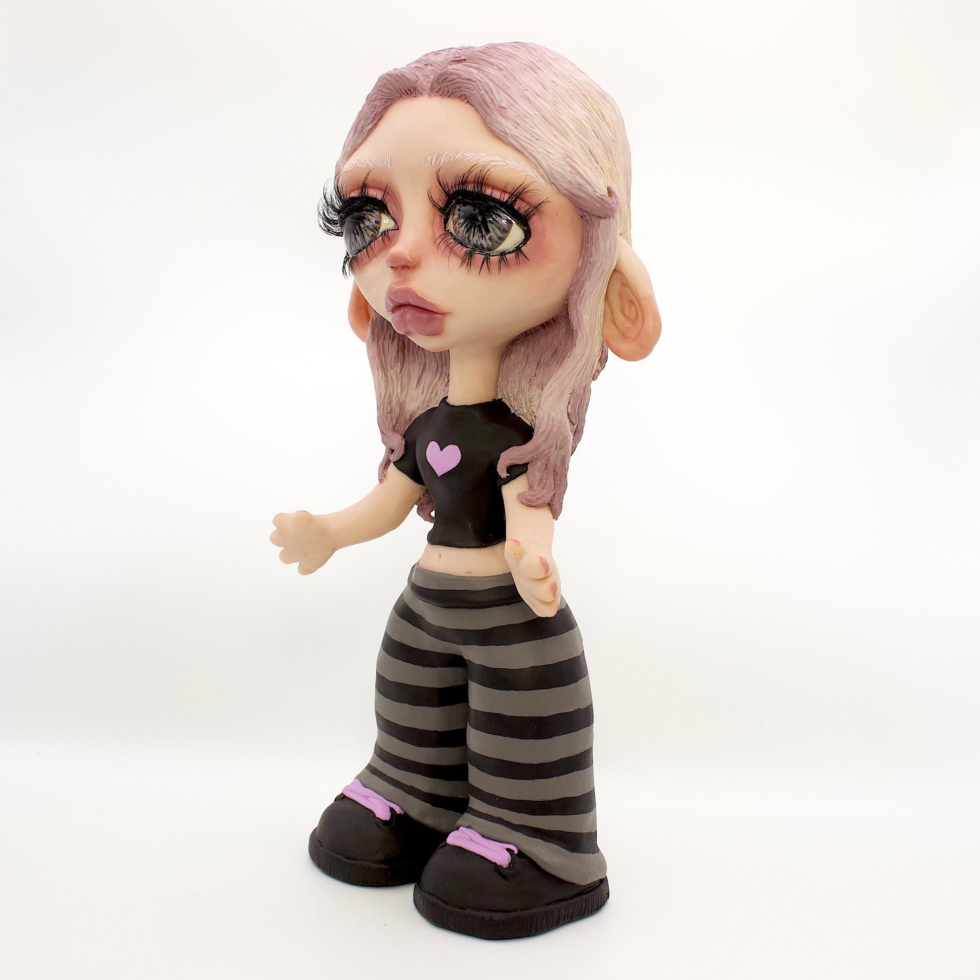 A stylized doll or figurine with large eyes, pale skin, lavender hair, and a black top with a pink heart, paired with black-and-gray striped pants and black shoes, photographed against a white studio backdrop.