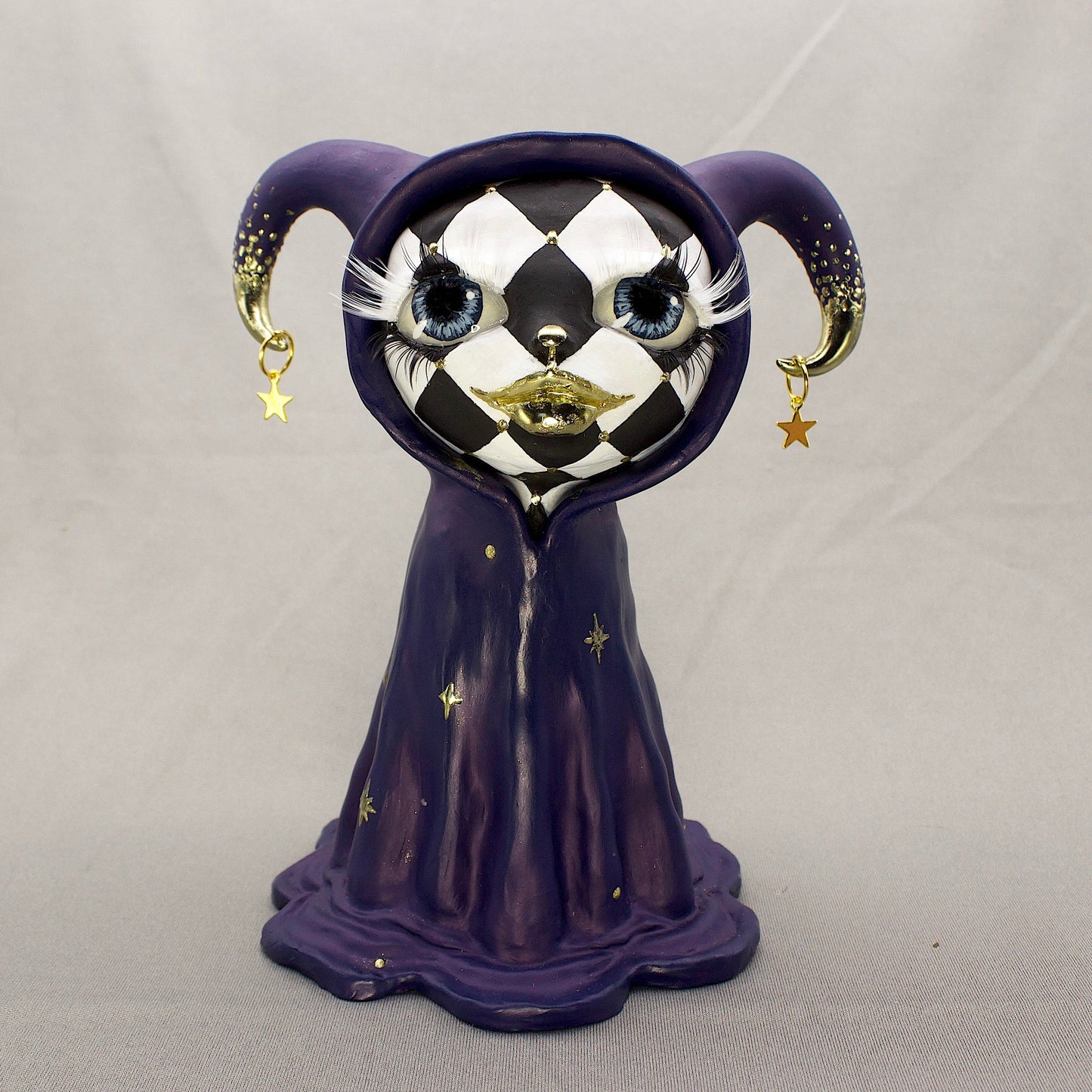 A black-and-white harlequin-pattern polymer clay figurine in a blue robe with gold accents, displayed on a light gray fabric backdrop.
