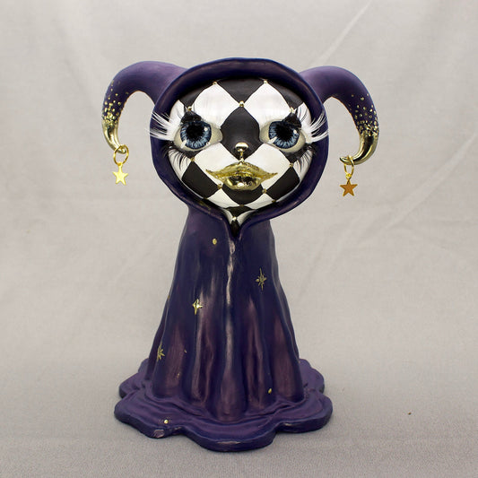A black-and-white harlequin-pattern polymer clay figurine in a blue robe with gold accents, displayed on a light gray fabric backdrop.