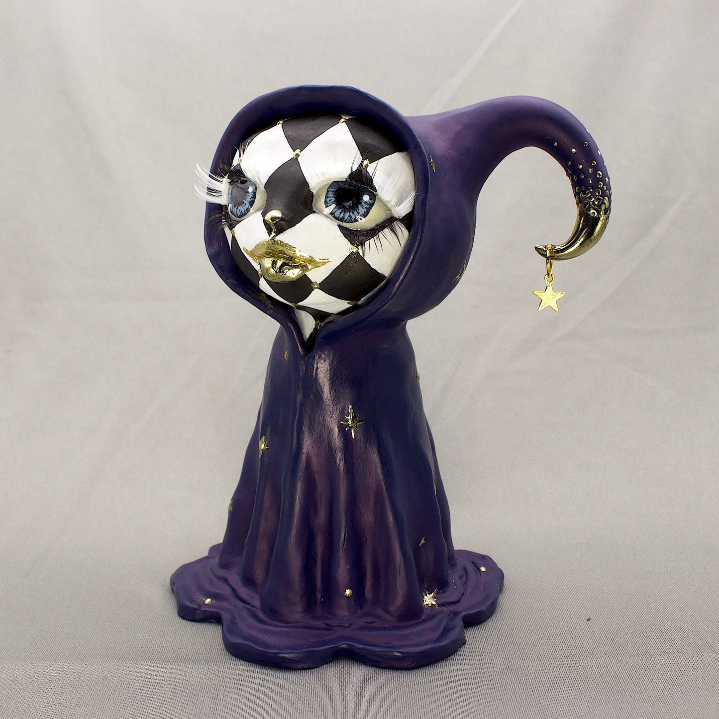 A black-and-white harlequin-pattern polymer clay figurine in a blue robe with gold accents, displayed on a light gray fabric backdrop.