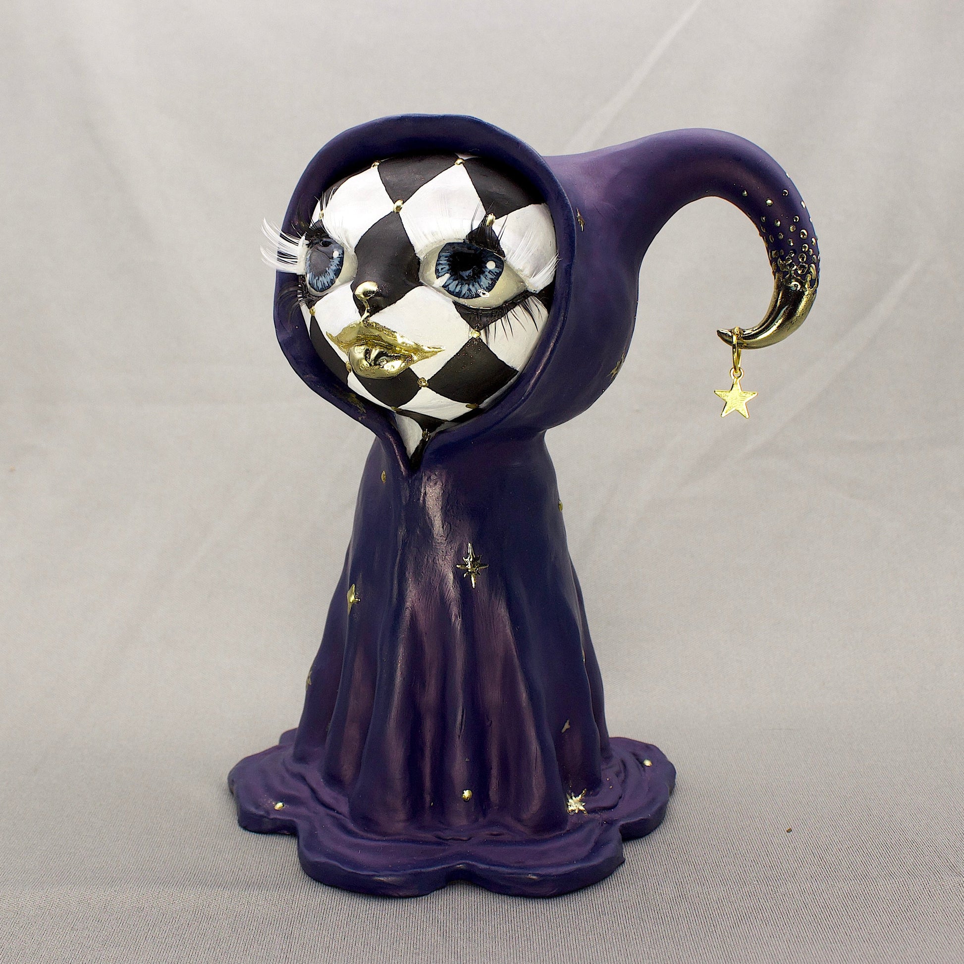 A black-and-white harlequin-pattern polymer clay figurine in a blue robe with gold accents, displayed on a light gray fabric backdrop.