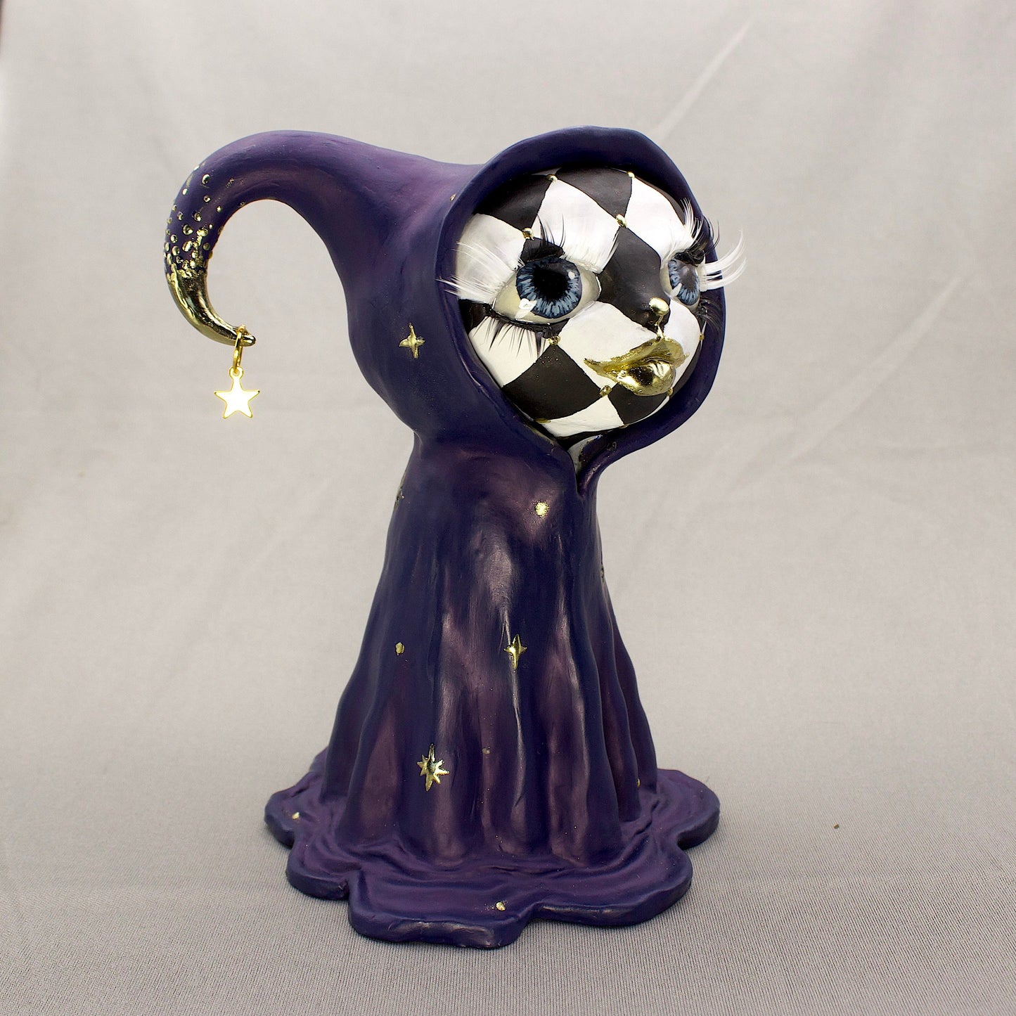 A black-and-white harlequin-pattern polymer clay figurine in a blue robe with gold accents, displayed on a light gray fabric backdrop.