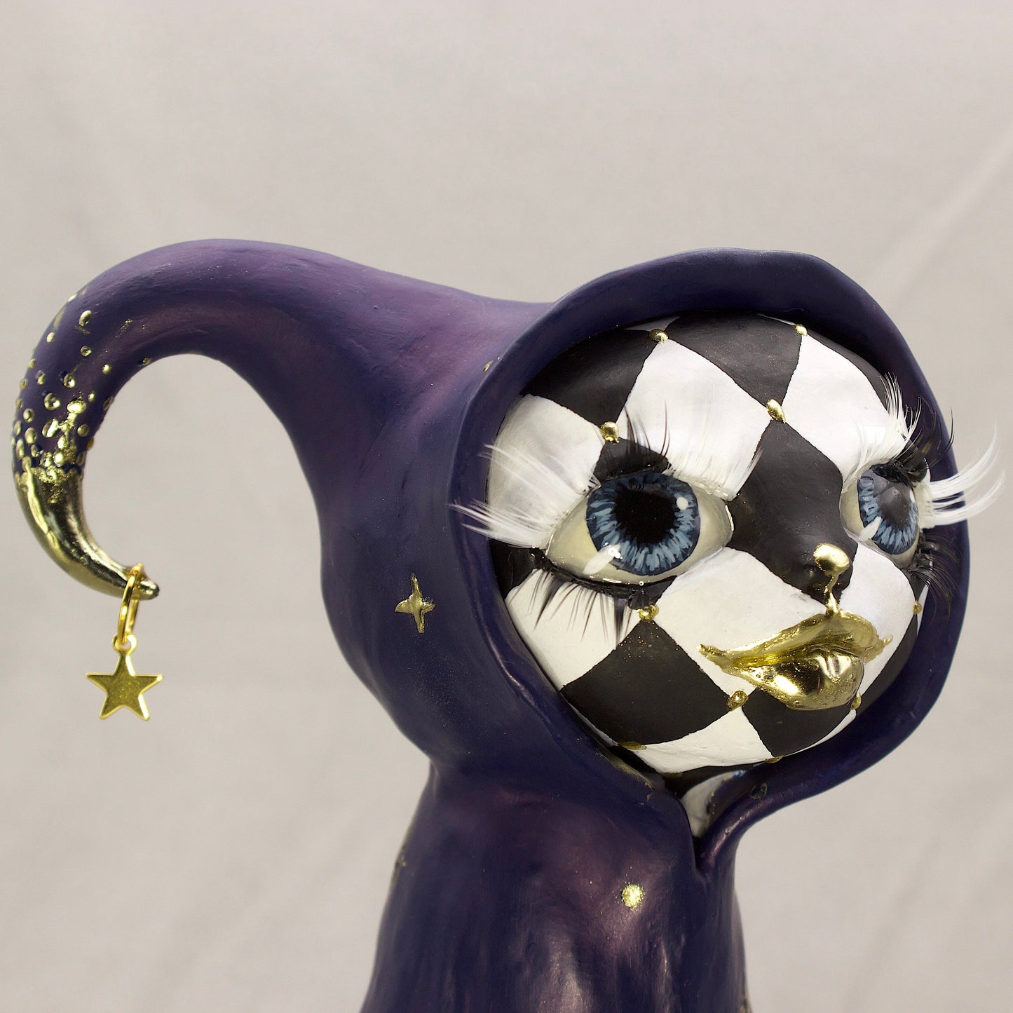 A black-and-white harlequin-pattern polymer clay figurine in a blue robe with gold accents, displayed on a light gray fabric backdrop.