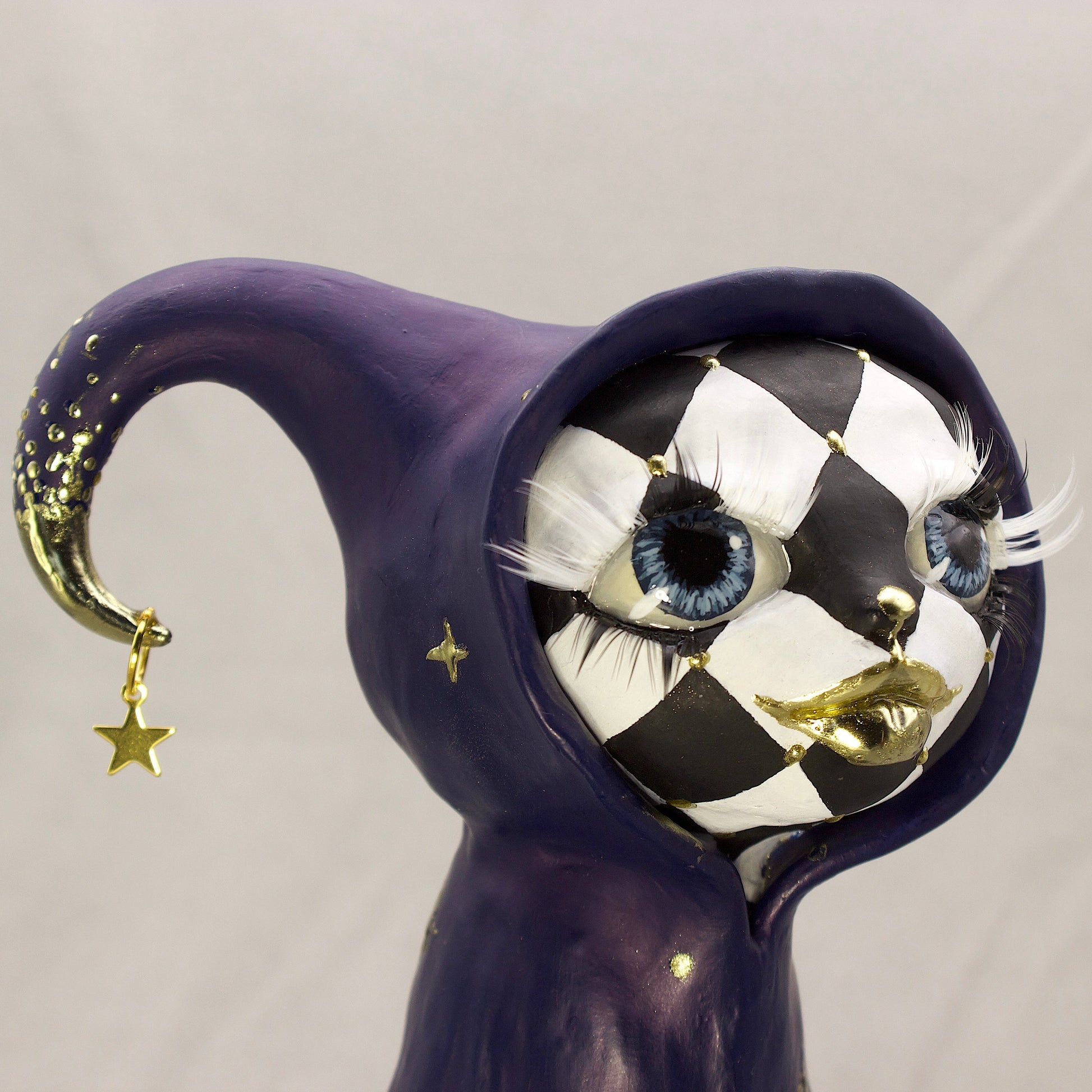 A black-and-white harlequin-pattern polymer clay figurine in a blue robe with gold accents, displayed on a light gray fabric backdrop.