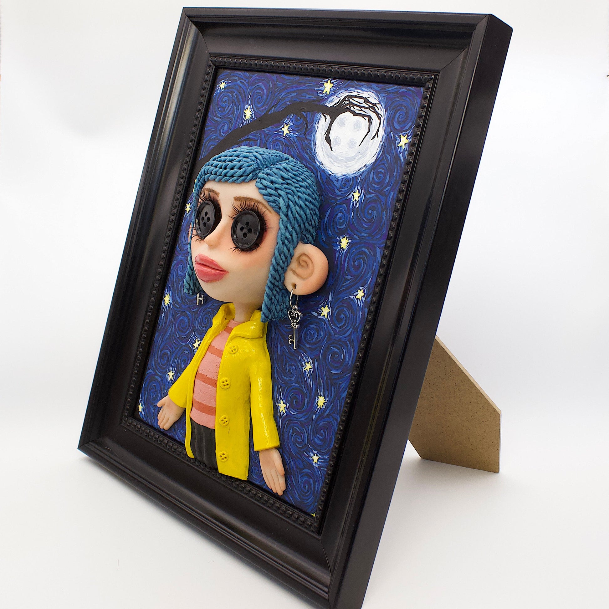 A polymer clay sculpture positioned in a black wood photo frame. Featuring the Coraline doll with her iconic button eyes, in her yellow raincoat, set against an impressionistic view of the starry sky with the button moon and a gnarled tree branch in the shape of the other mothers hand. 