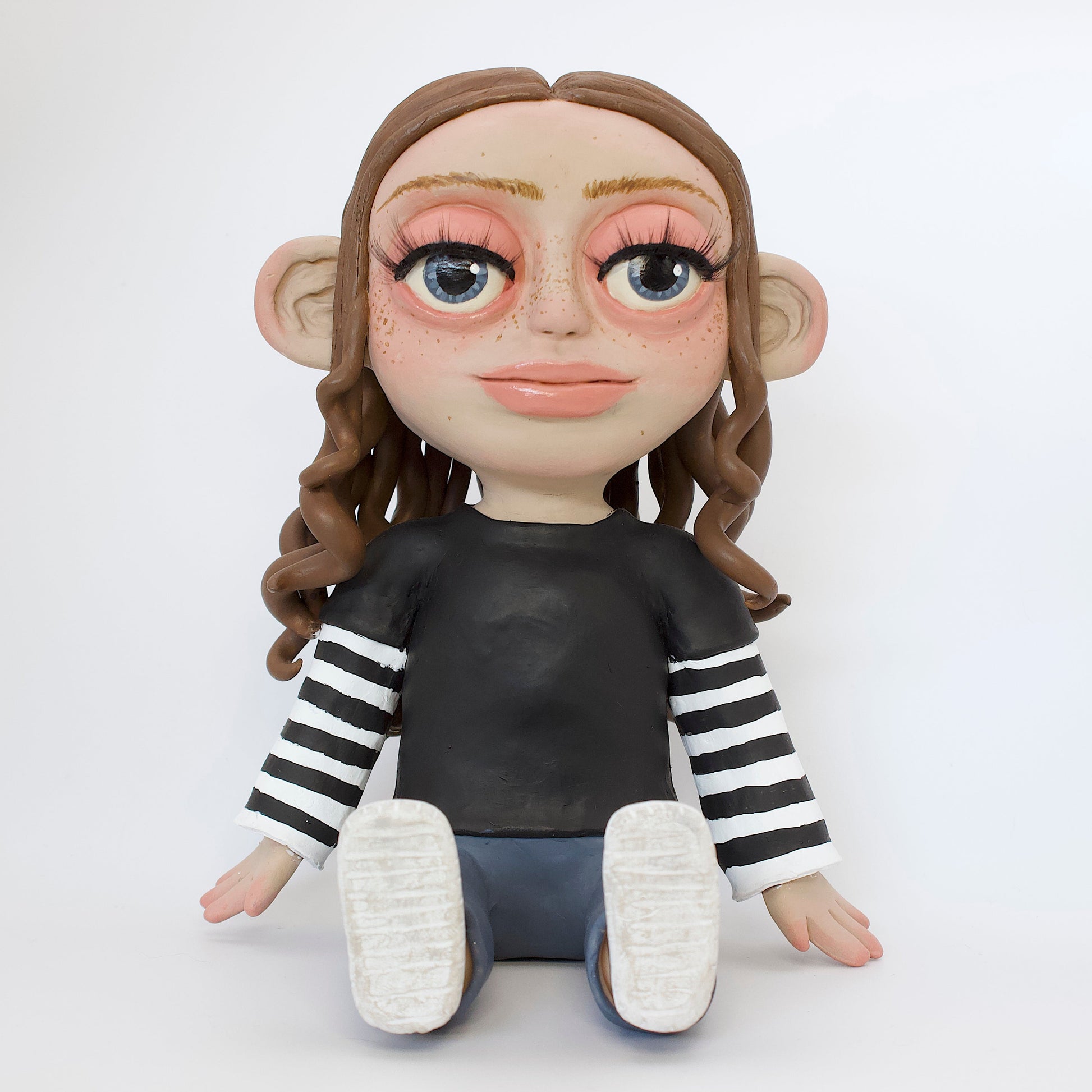 Polymer clay sculpture of a girl sitting down, featuring brown curly hair and skater style clothes, a black tshirt over a black and white striped long sleeve, with blue jeans and converse. 