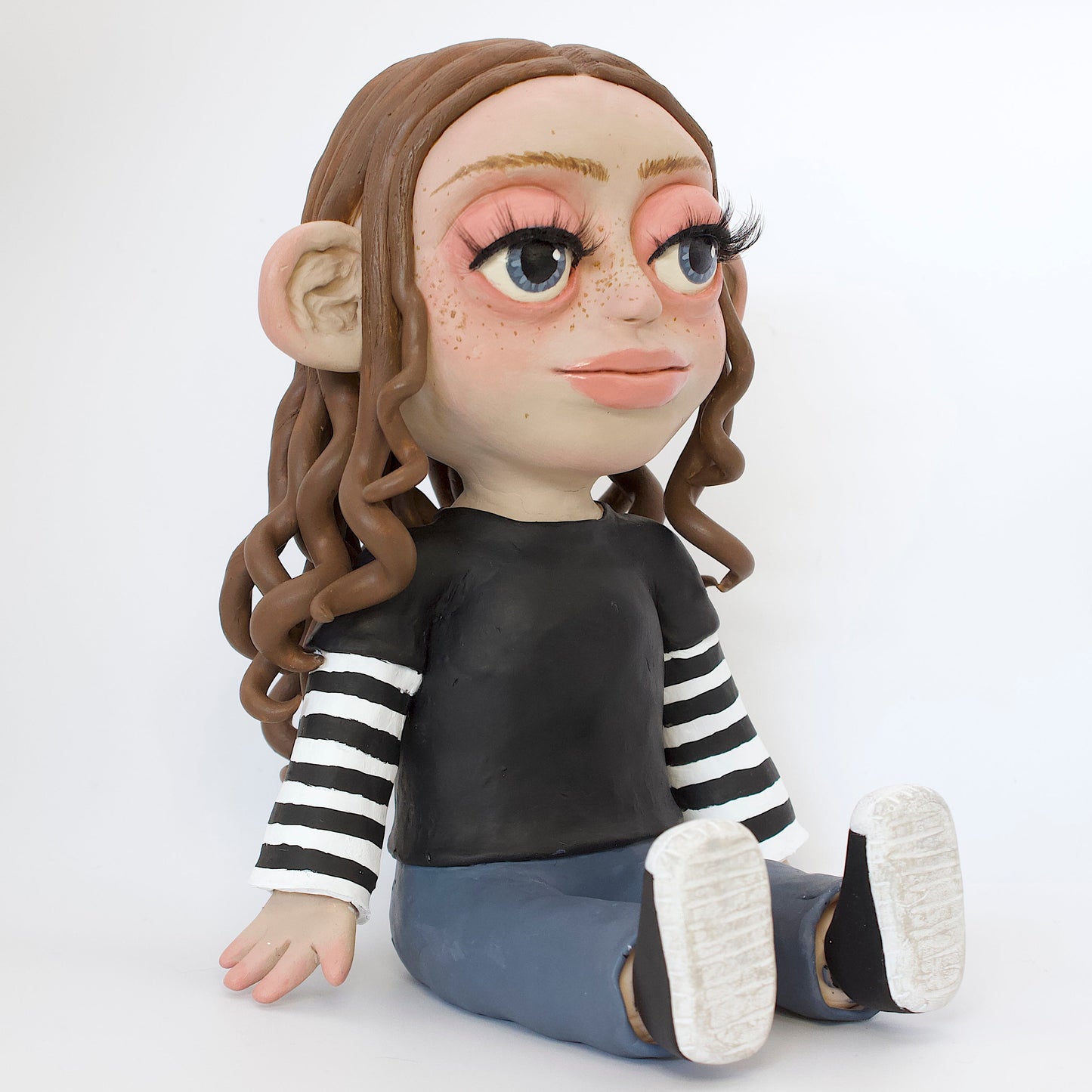Polymer clay sculpture of a girl sitting down, featuring brown curly hair and skater style clothes, a black tshirt over a black and white striped long sleeve, with blue jeans and converse. 