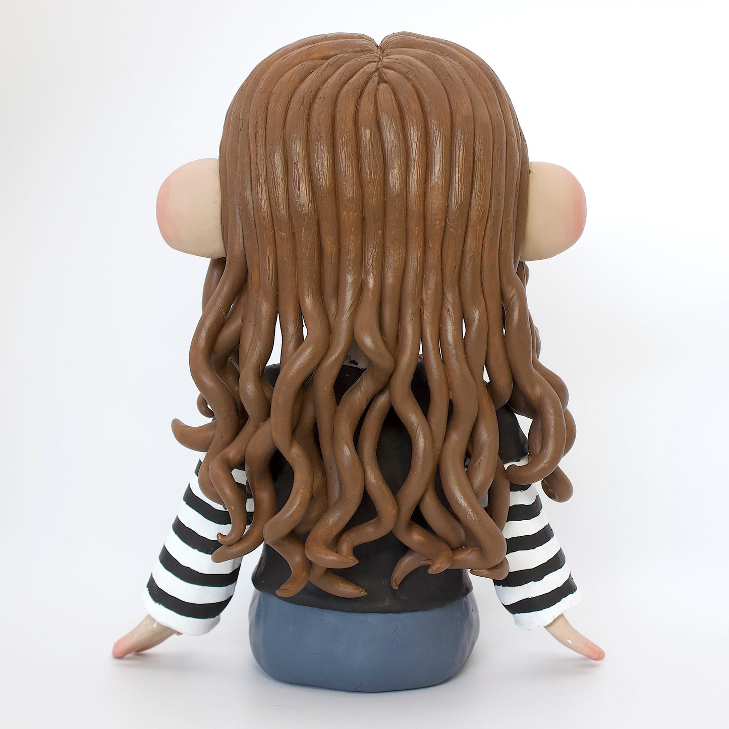 Polymer clay sculpture of a girl sitting down, featuring brown curly hair and skater style clothes, a black tshirt over a black and white striped long sleeve, with blue jeans and converse. 