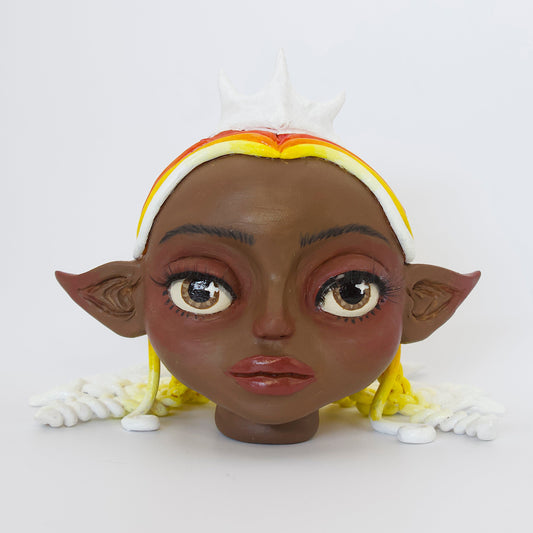 Polymer clay sculpture of a sun elf head, featuring a sun shaped crown, braided hair in a gradient from red, to orange, to yellow, to white. 