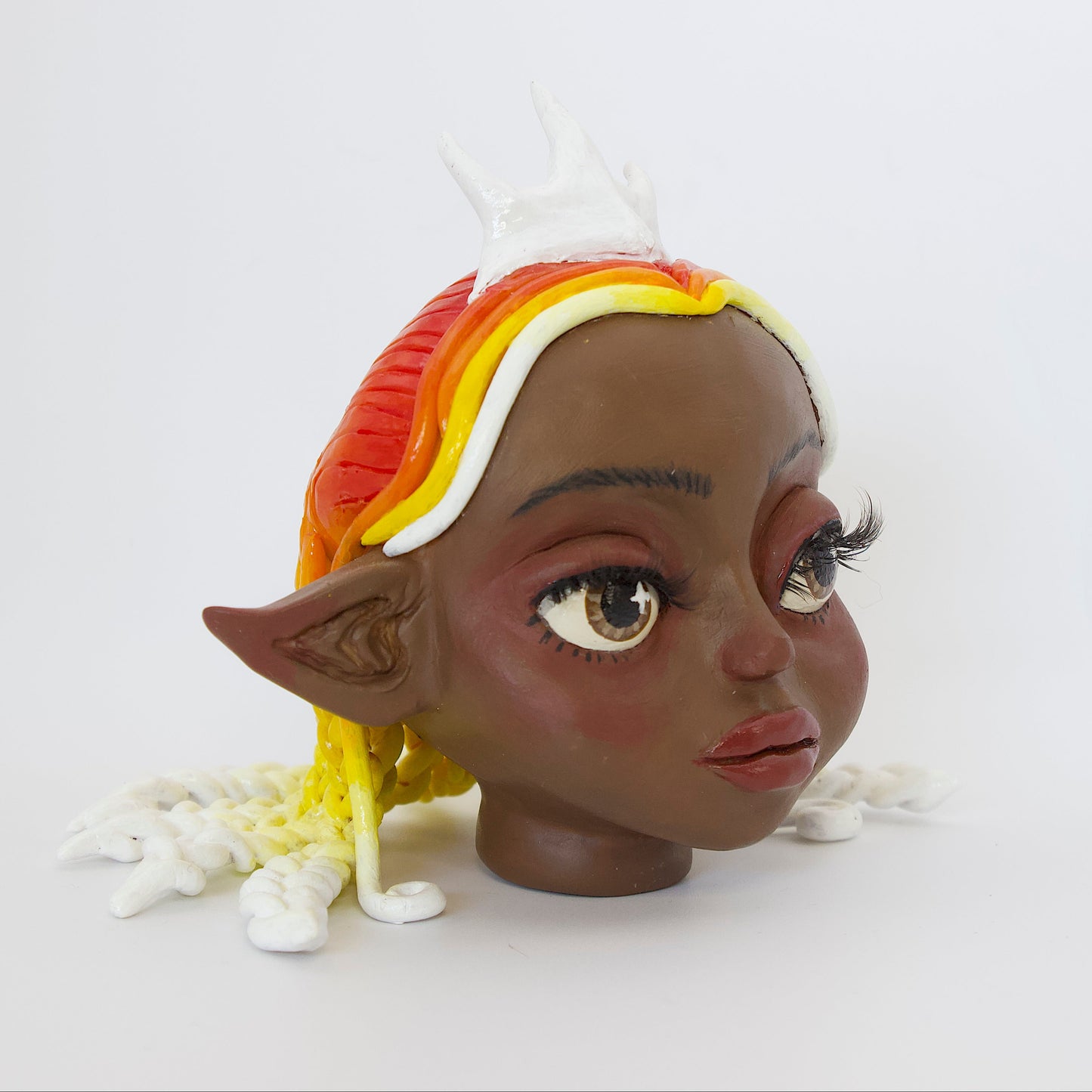 Polymer clay sculpture of a sun elf head, featuring a sun shaped crown, braided hair in a gradient from red, to orange, to yellow, to white. 